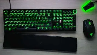 A Razer Huntsman V3 Pro seen top-down on a wooden surface alongside a mouse and mouse dock, with the wrist rest slightly apart